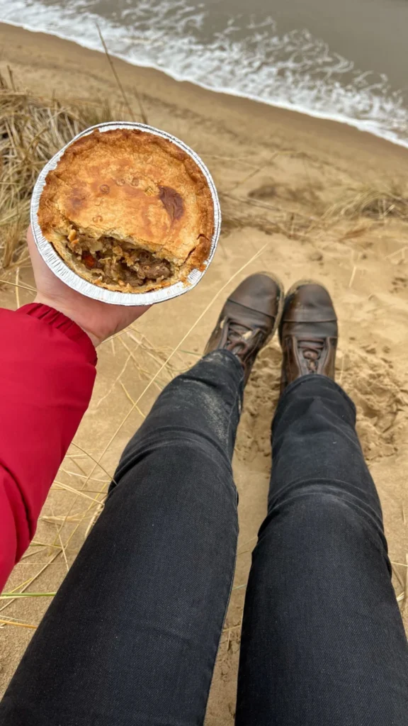 Ever Prel holding a partially eaten homebaked Anfield pie on Formby Beach with sand and sea in the background