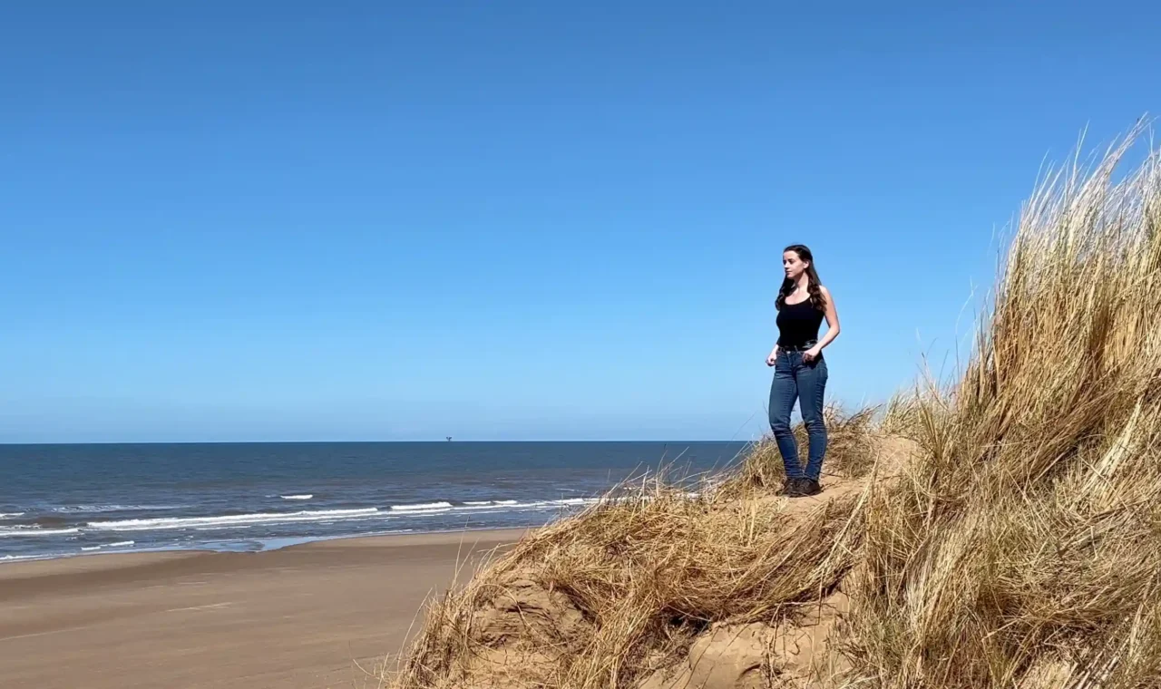 A young girl named Ever Prel standing on top of a sand dune at Formby Beach, Liverpool, looking out at the Irish Sea on a sunny day.