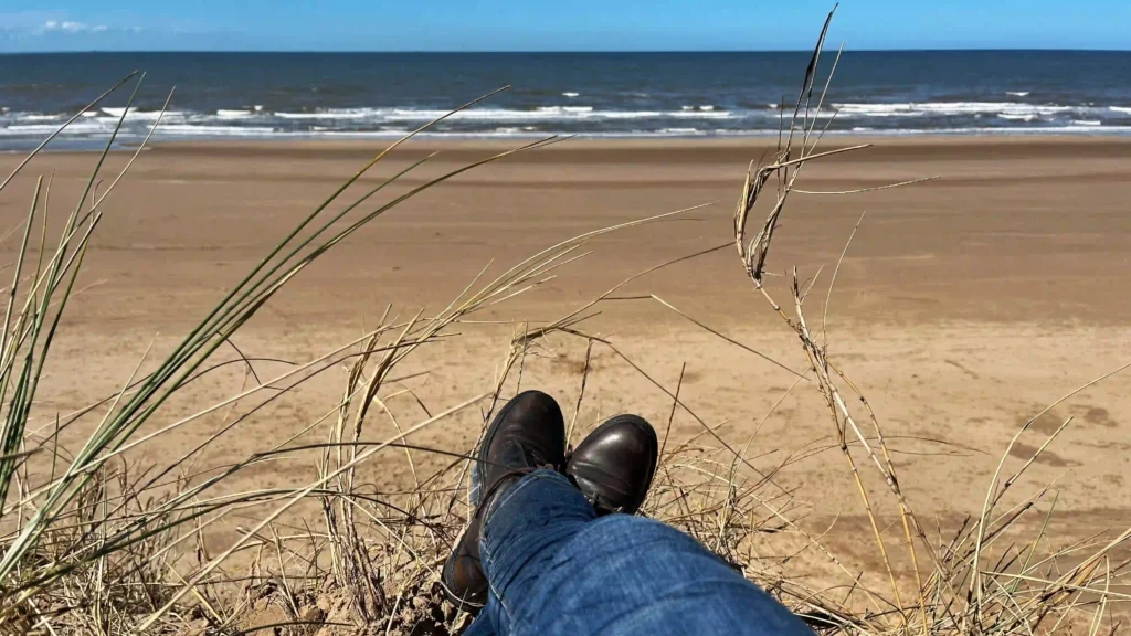Person sitting on a sand dune at Formby Beach, looking out at the Irish Sea on a sunny day.