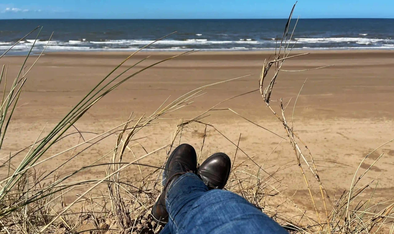 Person sitting on a sand dune at Formby Beach, looking out at the Irish Sea on a sunny day.