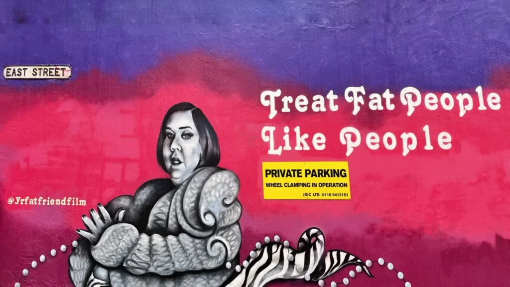 Street art mural in Nottingham, England, featuring a monochrome portrait of a stylish woman with the message “Treat fat people like people” on a vibrant purple and pink wall.