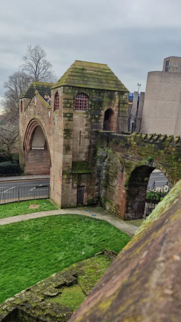 Chester City Walls in England supported by historic stone arcades, with a curved medieval walkway, square tower, park views and a modern road passing beneath.