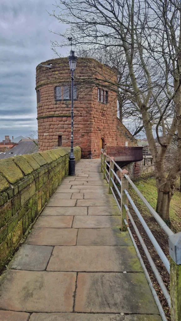 Historic Chester City Walls in England, featuring a red sandstone medieval walkway, moss-covered stone walls, a defensive tower and views over the rooftops of Chester.