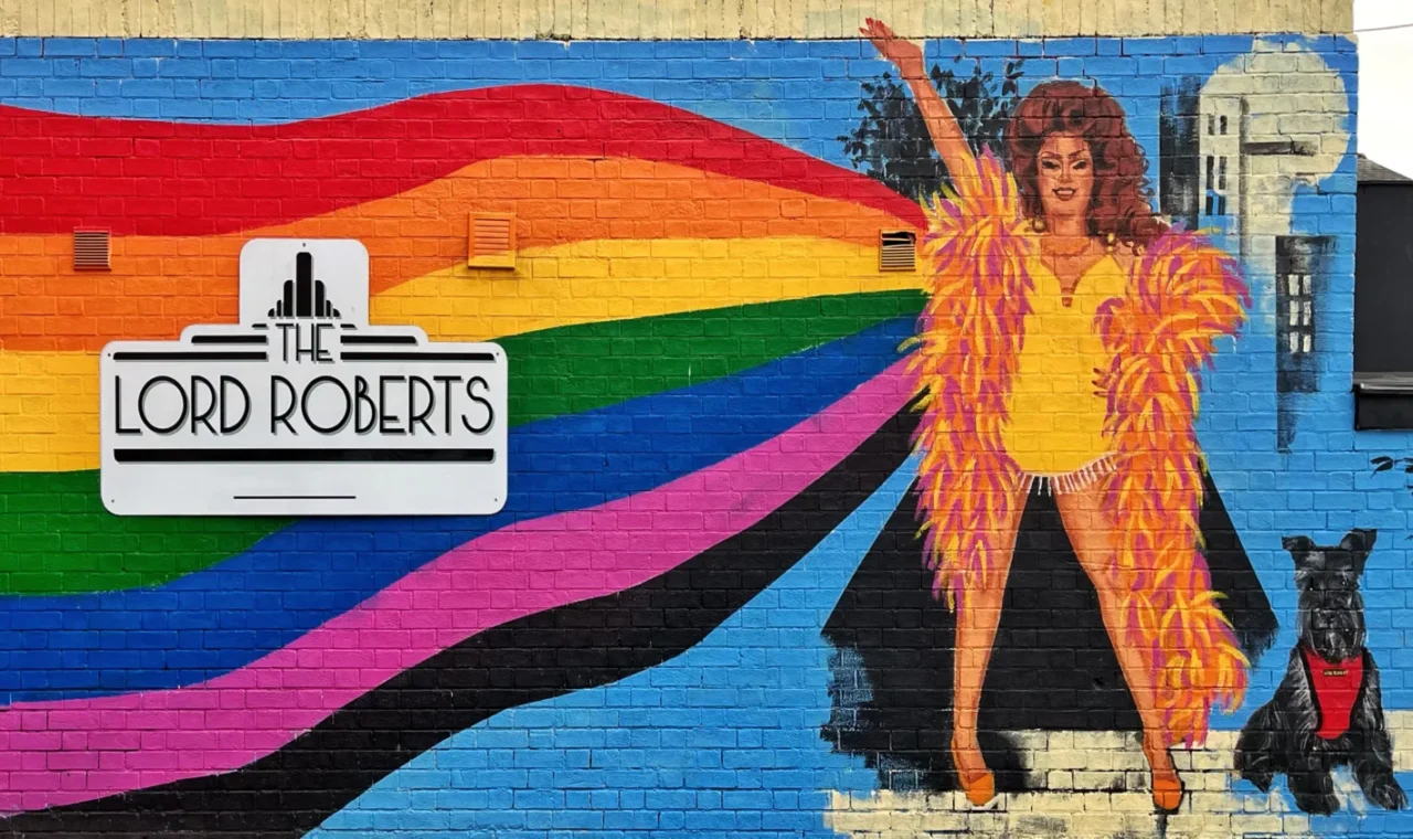 Colourful street art mural in Nottingham, England, depicting a vintage-inspired female performer with a dog beneath a rainbow against a blue cityscape background.