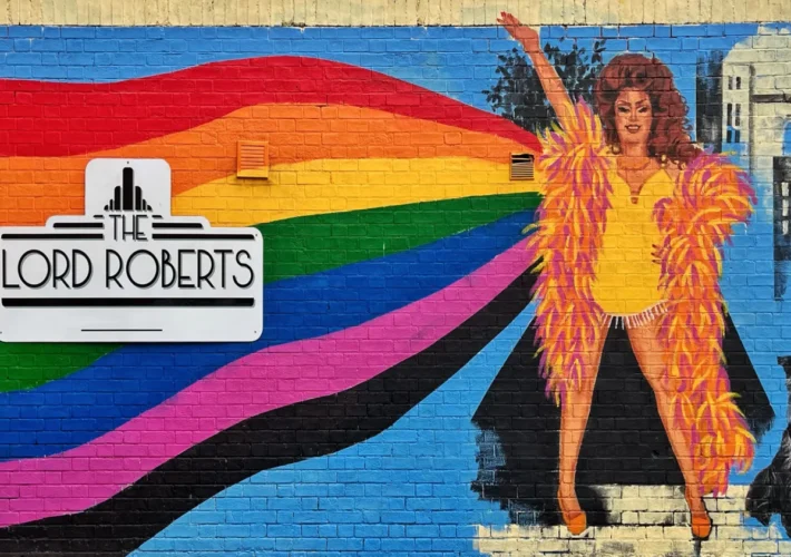 Colourful street art mural in Nottingham, England, depicting a vintage-inspired female performer with a dog beneath a rainbow against a blue cityscape background.