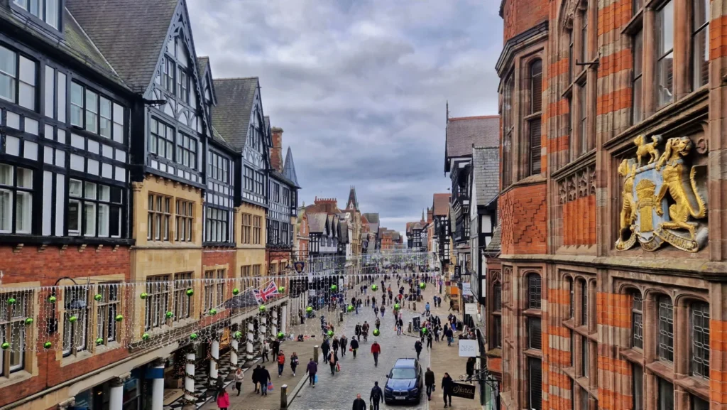 View along Eastgate Street in Chester, England, from Chester Cross, featuring the Eastgate Clock, historic half-timbered buildings, Christmas decorations and visitors walking through the city centre.