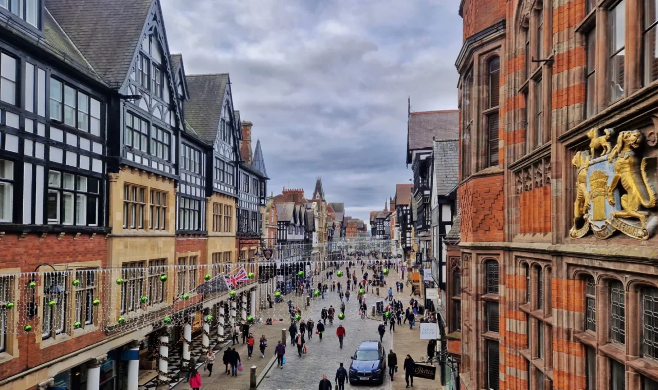 View along Eastgate Street in Chester, England, from Chester Cross, featuring the Eastgate Clock, historic half-timbered buildings, Christmas decorations and visitors walking through the city centre.