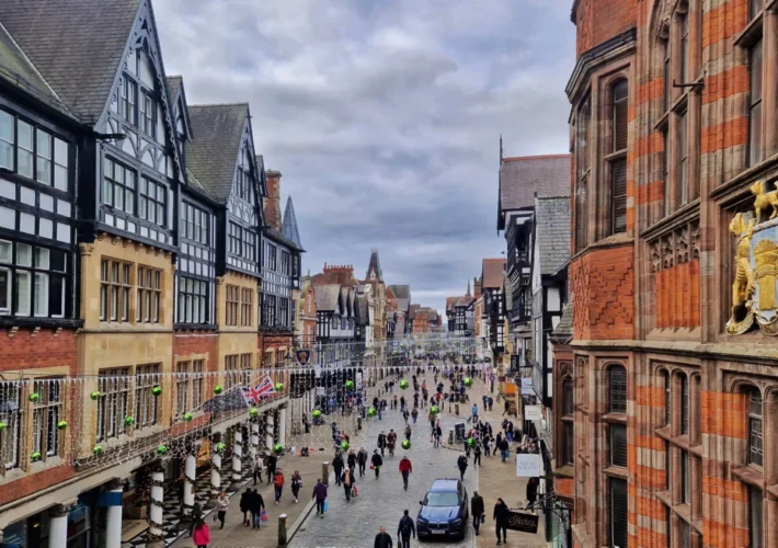 View along Eastgate Street in Chester, England, from Chester Cross, featuring the Eastgate Clock, historic half-timbered buildings, Christmas decorations and visitors walking through the city centre.
