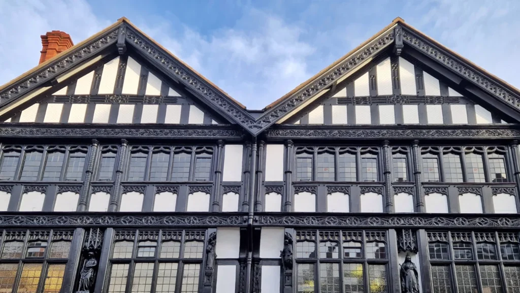 Decorative black-and-white timber-framed gabled houses in Chester, England, featuring intricate wood carvings, stained-glass windows, carved wooden figures and a red brick chimney beneath a blue evening sky.