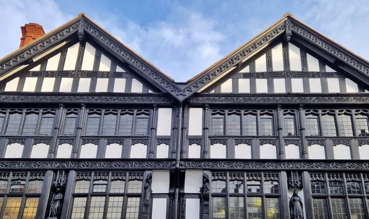 Decorative black-and-white timber-framed gabled houses in Chester, England, featuring intricate wood carvings, stained-glass windows, carved wooden figures and a red brick chimney beneath a blue evening sky.