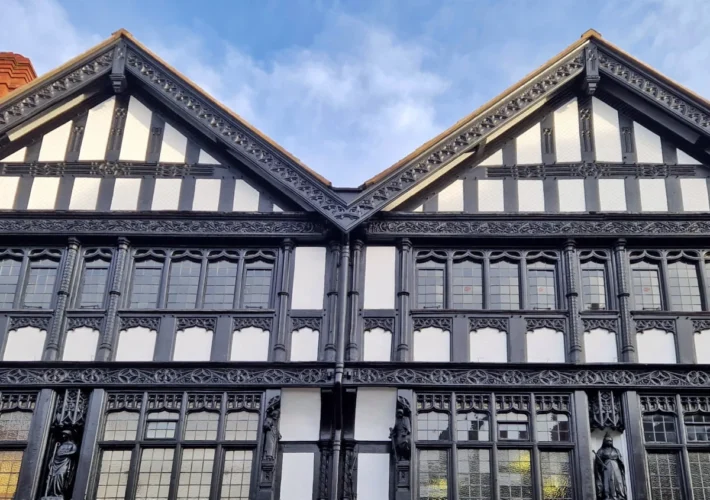 Decorative black-and-white timber-framed gabled houses in Chester, England, featuring intricate wood carvings, stained-glass windows, carved wooden figures and a red brick chimney beneath a blue evening sky.
