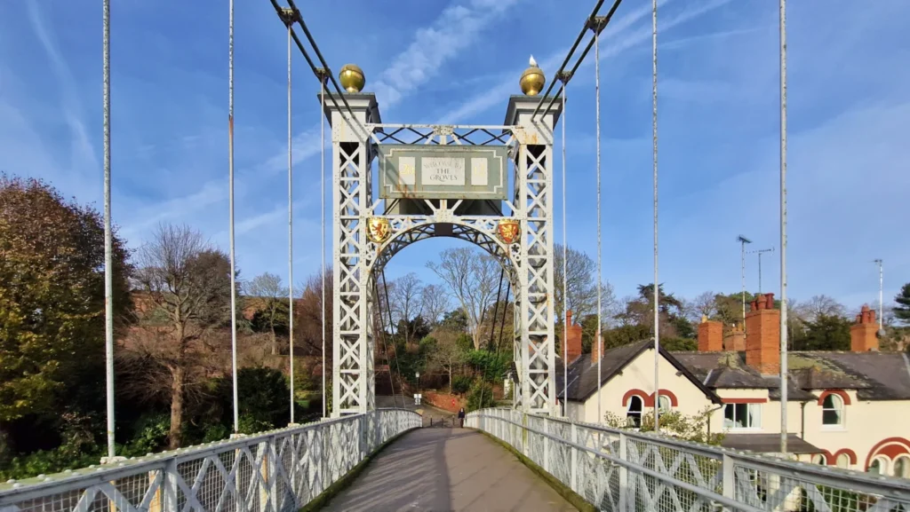 Queens Park Suspension Bridge in Chester, England, viewed in winter beneath a blue sky, featuring its Victorian iron structure, the “Welcome to The Groves” sign and decorative coats of arms.