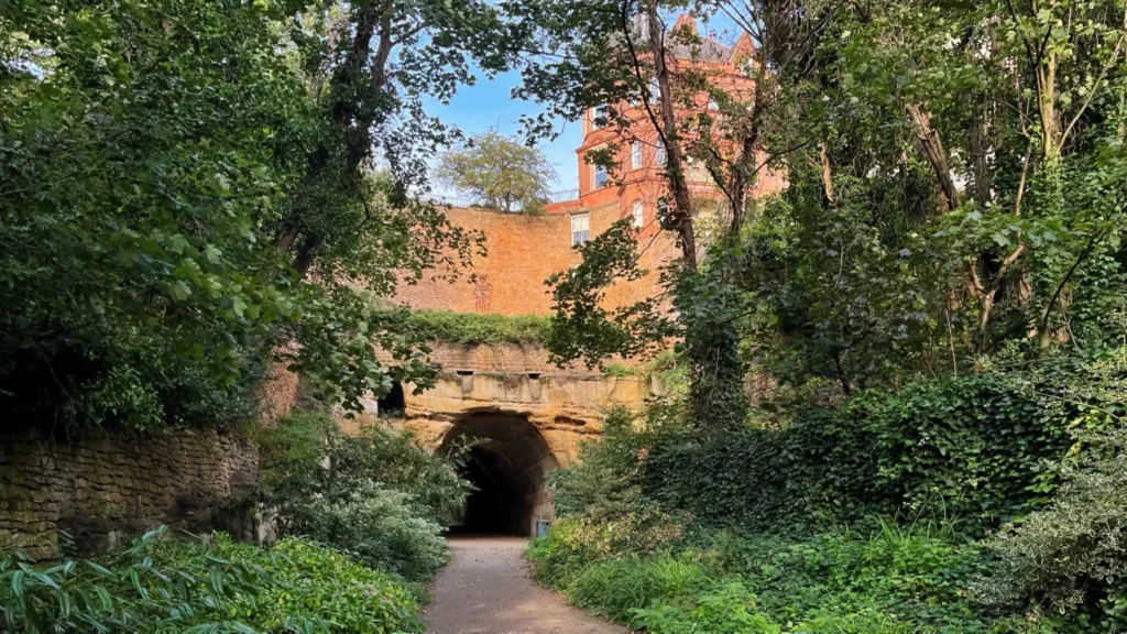 Woodland path leading through a historic rock tunnel called Park Tunnel (one of many Nottingham hidden gems) beneath Nottingham Castle in Nottingham, England, surrounded by lush greenery and overlooked by the castle.