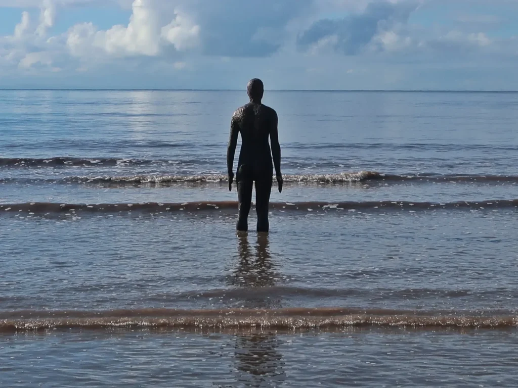 Cast-iron figure from Antony Gormley's Another Place installation standing in the shallow tidal waters of Crosby Beach, Merseyside, facing the horizon under a partly cloudy sky