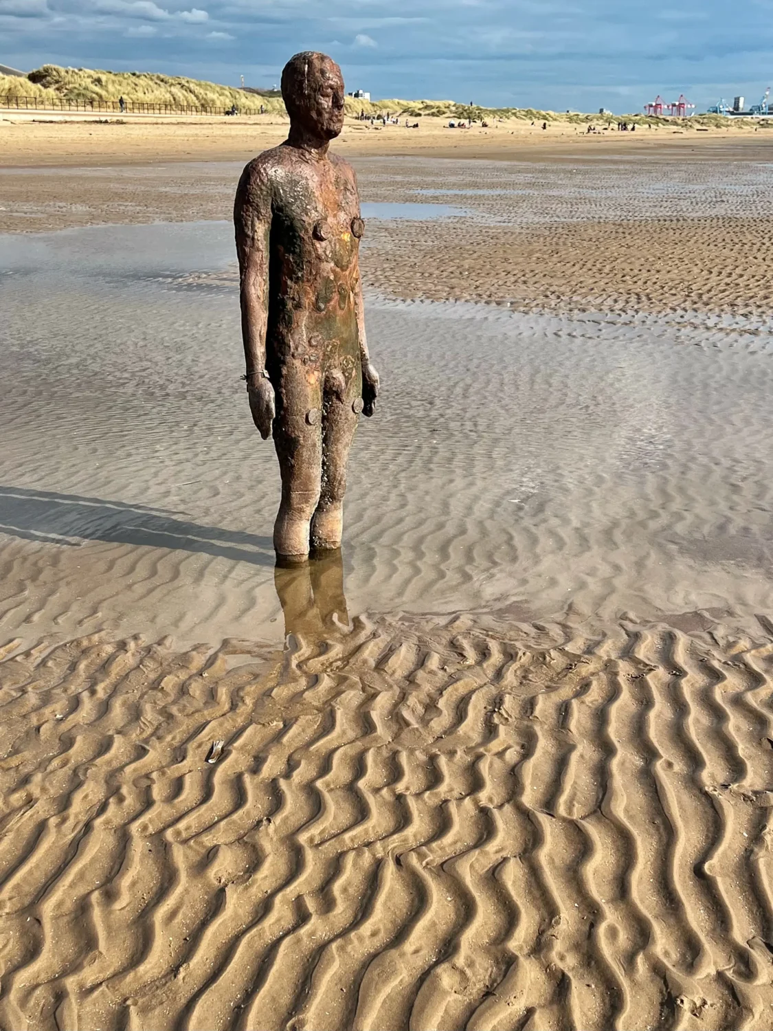 One of Antony Gormley's cast-iron Another Place figures standing in the sand at Crosby Beach, tide out, overcast sky