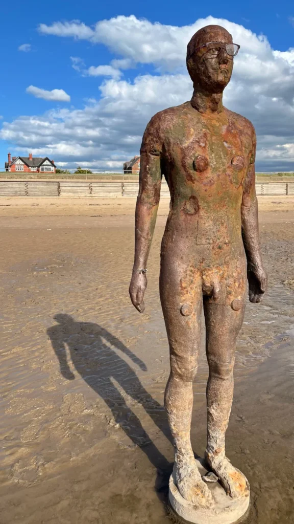 Full-length view of one of Antony Gormley's cast-iron figures, wearing glasses, standing on the sand at Crosby Beach