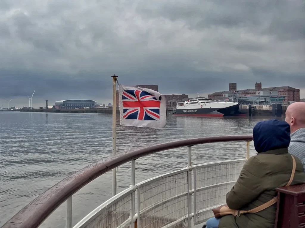 View from a Mersey Ferry deck with a Union Jack flag, passengers, and the Liverpool waterfront across the river